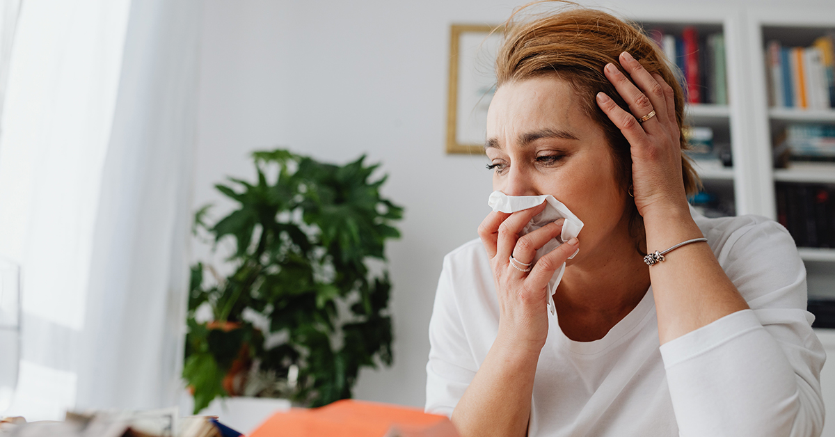 Woman covers nose and mouth with kleenex with hand on hand feeling sad